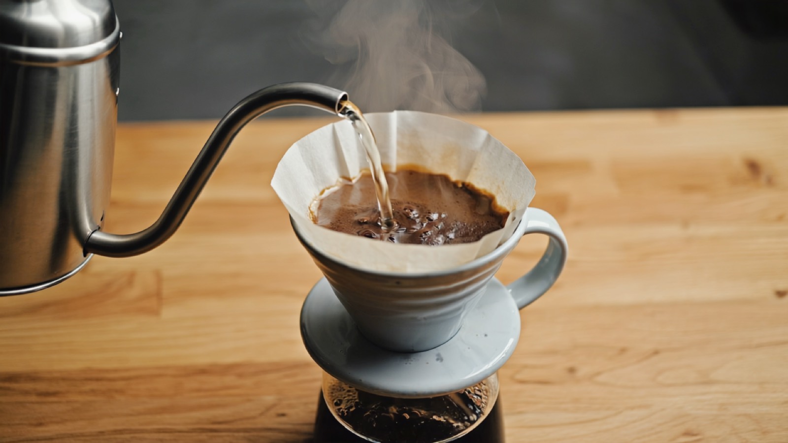 Heißes Wasser wird aus einem Schwanenhalskessel in einen Keramik-Handfilter mit Papierfilter gegossen, aufquellendes Kaffeemehl und aufsteigender Dampf im warmen Küchenlicht